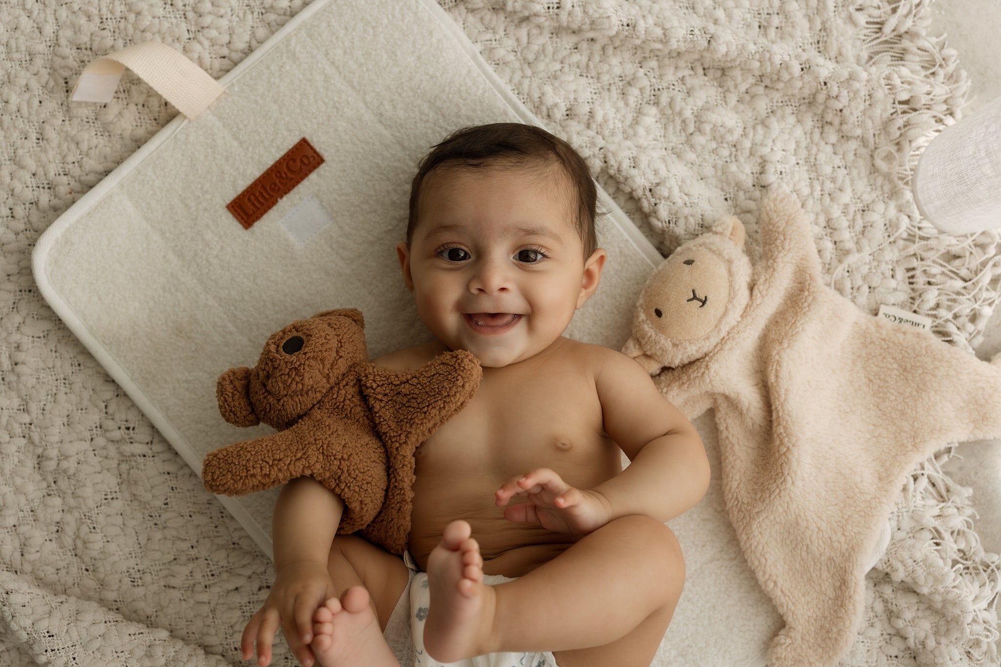 Baby lying on a rug with a teddy bear, smiling at the camera.