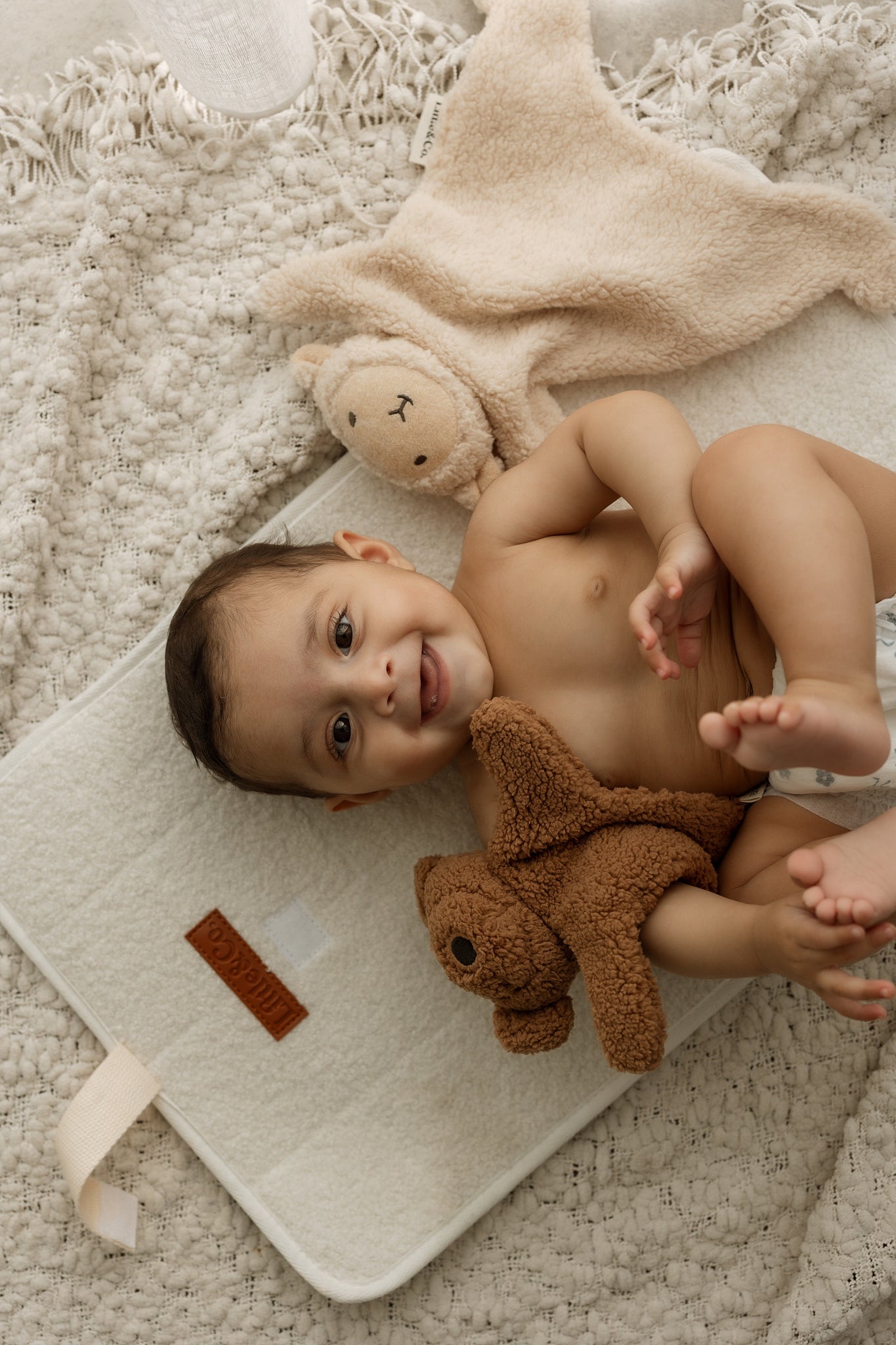 Baby lying on a rug with a teddy bear, smiling at the camera.