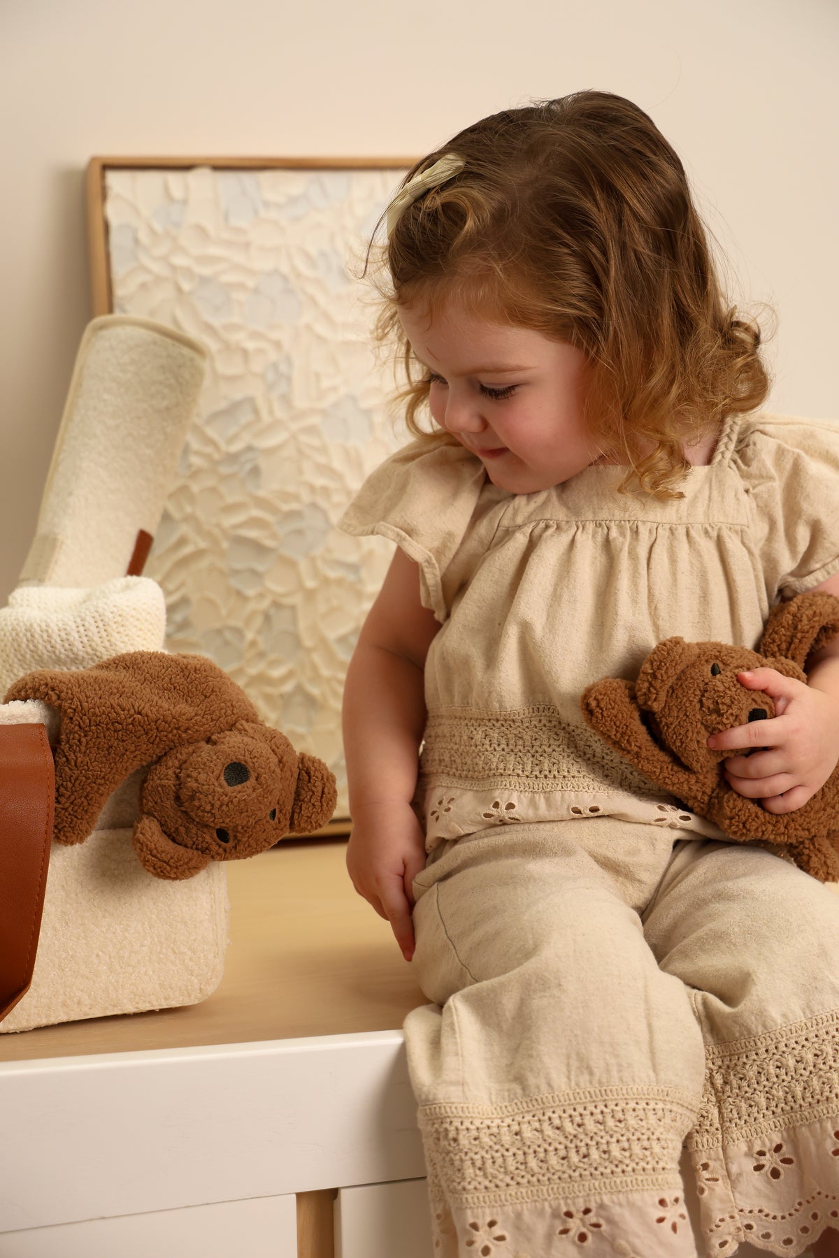 Toddler sitting on tall boy holding brown teddy teddy cuddly, soft neutral outfit and warm lighting