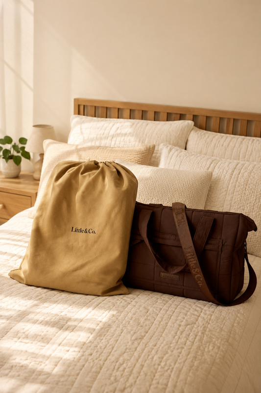 A brown duffel bag and a beige drawstring dust bag with 'Little&Co.' printed on it, styled on a bed with cream bedding and a wooden headboard in a warm, sunlit room.