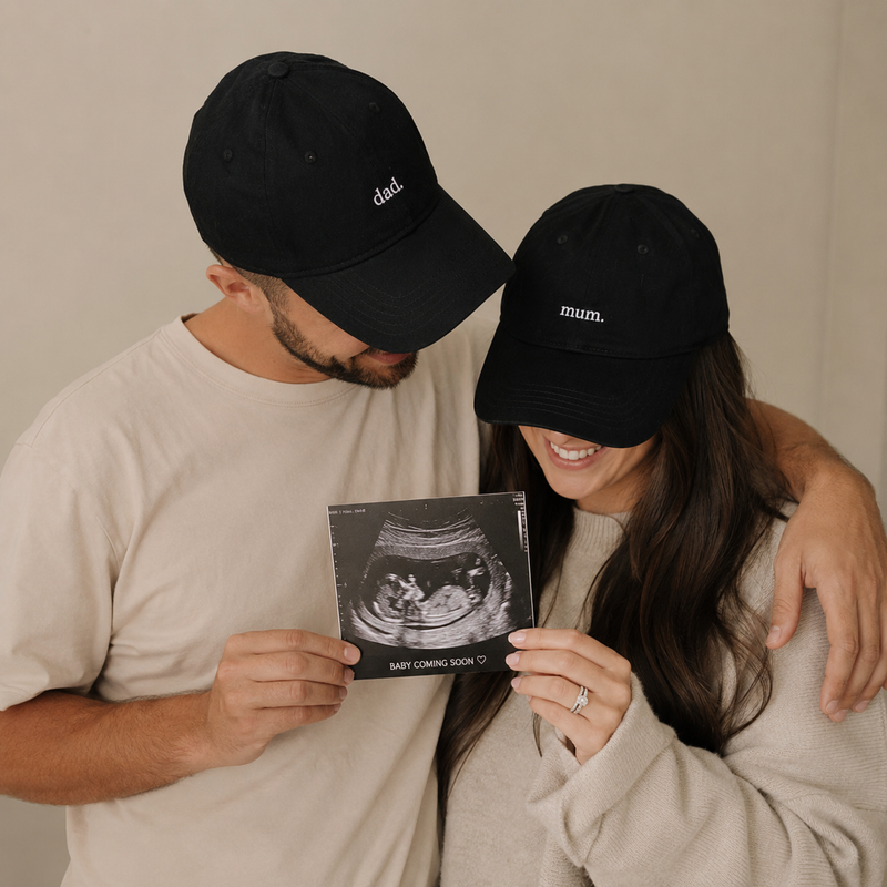 Man and woman wearing black caps with 'dad' and 'mum' text, holding an ultrasound photo together.