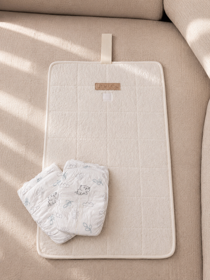 A cream-colored quilted baby changing pad with a small fabric brand label, shown on a beige upholstered sofa with two patterned baby diapers placed on top.