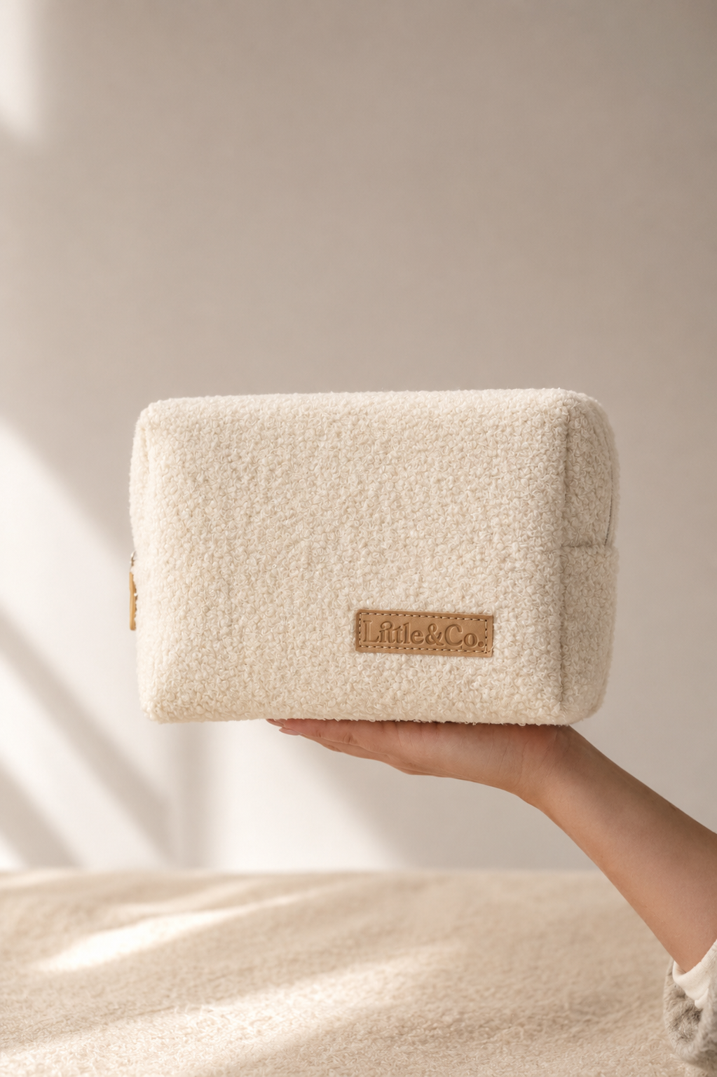 A cream-colored, textured bouclé pouch held by a hand against a warm beige background with soft window shadows.