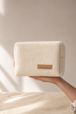A cream-colored, textured bouclé pouch held by a hand against a warm beige background with soft window shadows.