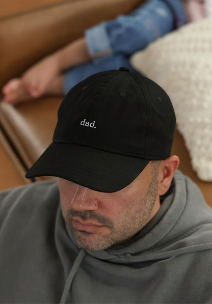 Man wearing a black cap with 'dad.' text, sitting on a couch.