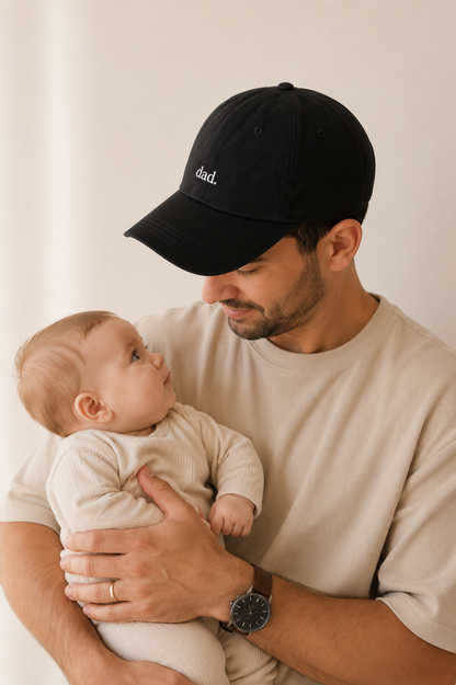 Man wearing a black cap with 'dad' embroidered on it, holding a baby against a plain background.