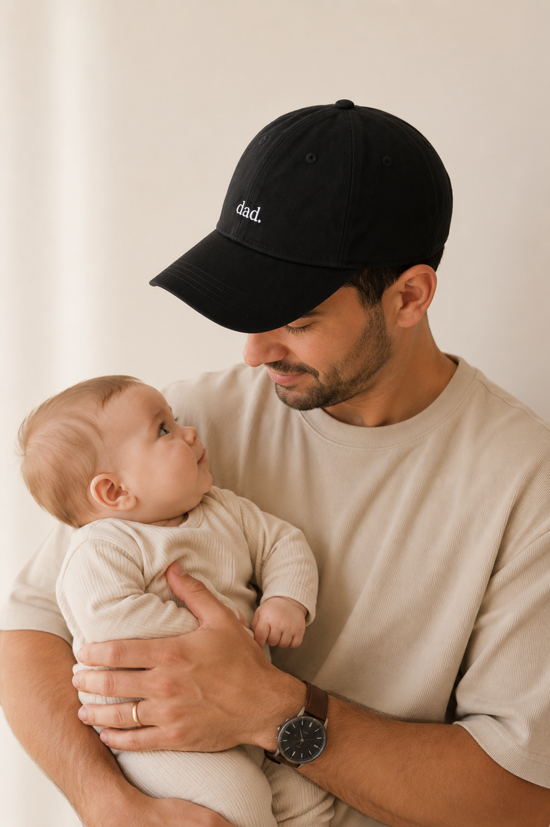 Man wearing a black cap with 'dad' embroidered on it, holding a baby against a plain background.