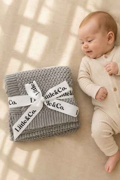 Baby lying next to a gray knitted blanket with 'Little & Co.' branding on a beige surface.