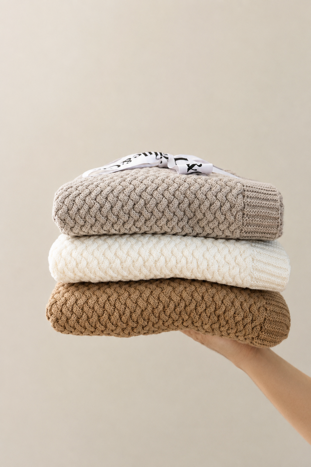 Stack of three knitted sweaters in beige, white, and brown held against a neutral background
