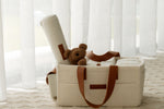 cream boucle nappy caddy with brown handles and a teddy bear inside, against a white curtain background.