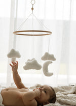 Baby reaching for a hanging cloud and moon mobile in a softly lit room.