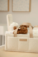 Beige bouclé nappy caddy with vanilla leather straps and logo, filled with baby essentials and teddy in neutral nursery setting
