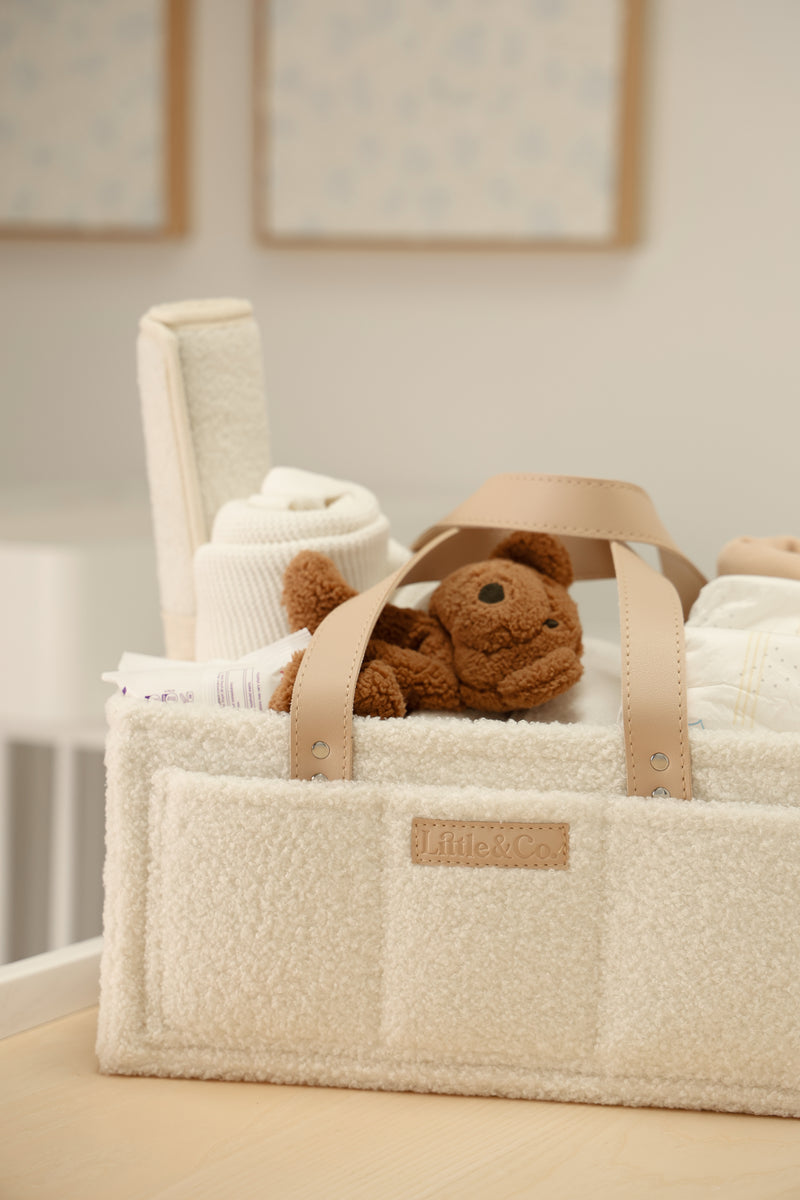 Beige bouclé nappy caddy with vanilla leather straps and logo, filled with baby essentials and teddy in neutral nursery setting