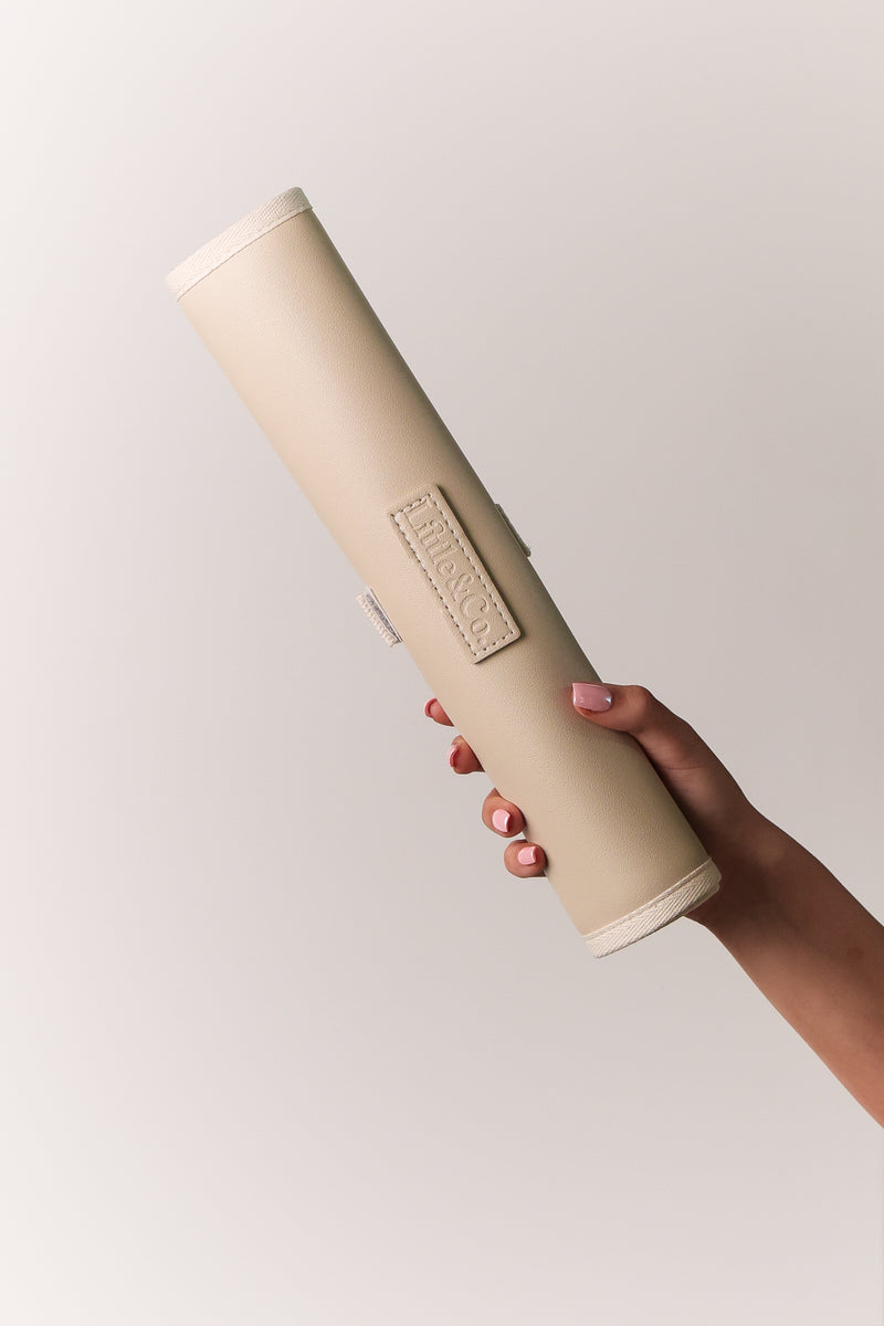 Hand holding a rolled beige leather baby change mat by Little & Co against a neutral background.