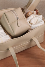 Beige leather baby nappy caddy filled with essentials including nappies, a zip pouch, and a rolled changing mat, styled on a wooden surface.
