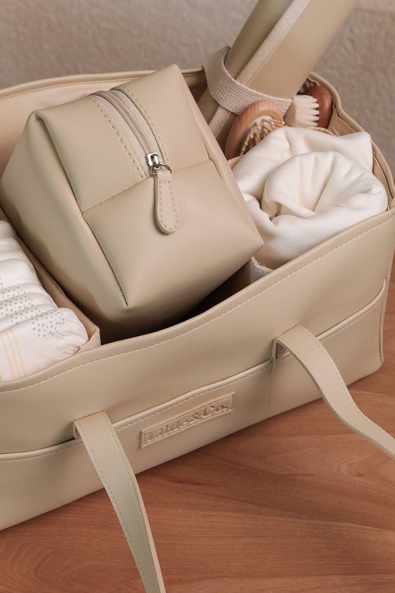 Beige leather baby nappy caddy filled with essentials including nappies, a zip pouch, and a rolled changing mat, styled on a wooden surface.
