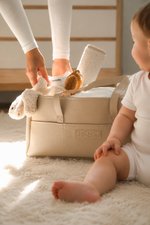 A beige baby diaper bag with a visible Little & Co brand patch sits on a white shag rug. A baby in a white onesie is sitting on the right, and a person's legs and hand are visible on the left placing a white plush toy into the bag.