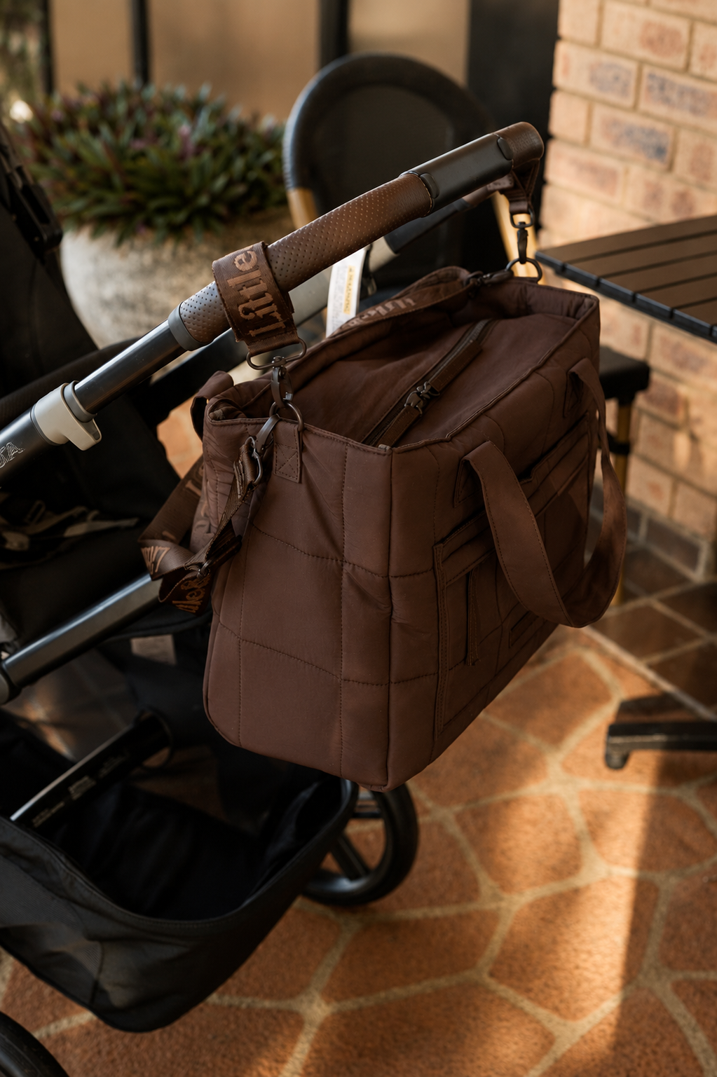 A brown diaper bag with quilted paneling and a top zipper, hanging from a black stroller handle in an outdoor patio setting.