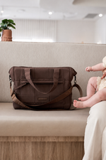 A brown padded diaper bag with a wide shoulder strap sits on a beige upholstered bench in a softly lit indoor setting. A baby's legs and feet are visible on the right side, and a small potted plant appears in the background.