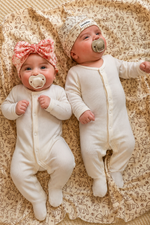 Two babies in white onesies with pacifiers lying on a patterned blanket.