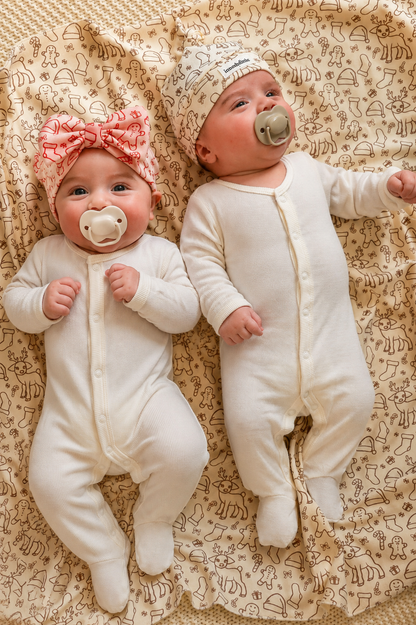 Two babies in white onesies with pacifiers lying on a patterned blanket.