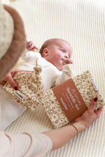 A baby swaddled in a beige blanket with a festive print, being held by a adult, with a beanie with a knot hat also visible.
