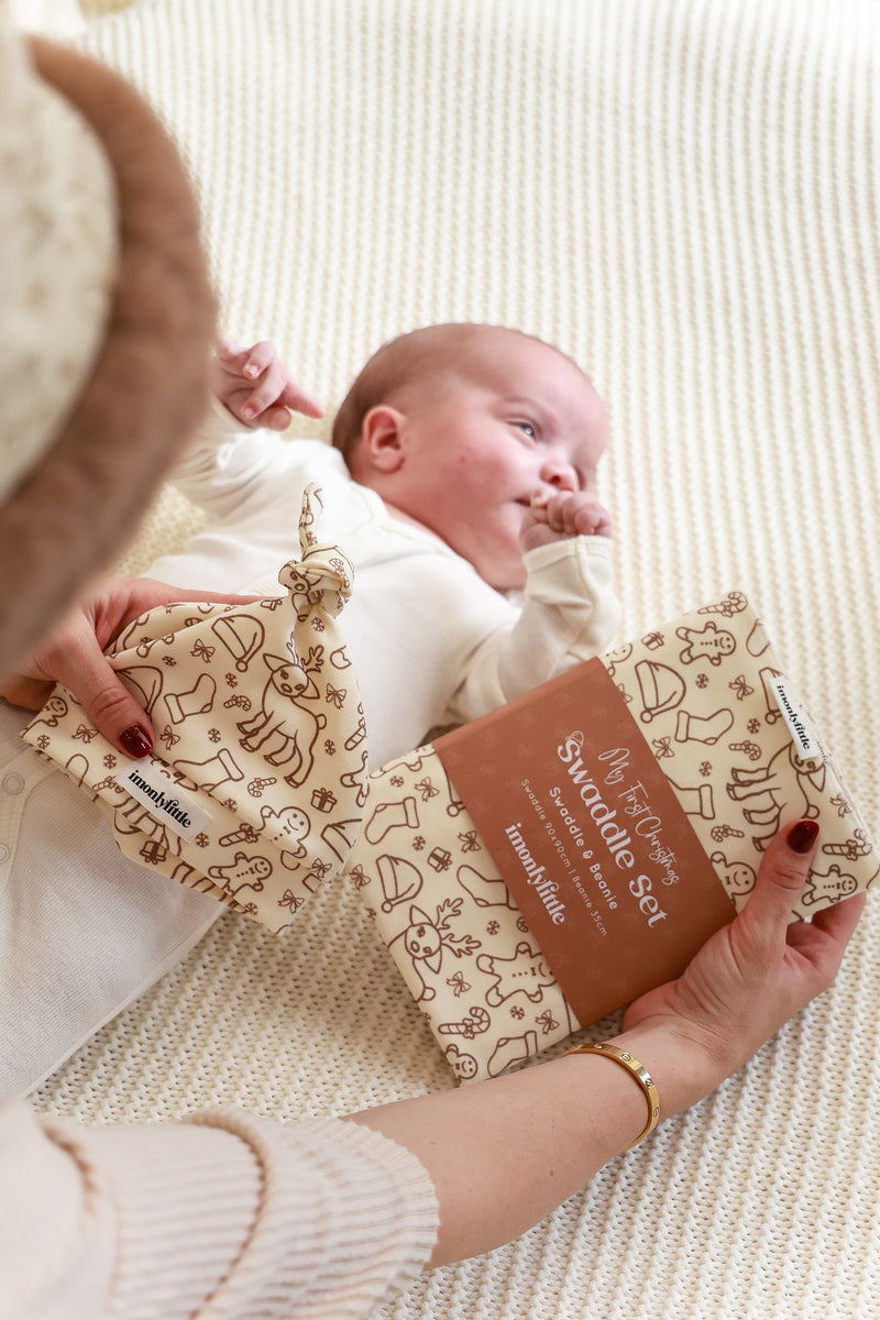 A baby swaddled in a beige blanket with a festive print, being held by a adult, with a beanie with a knot hat also visible.