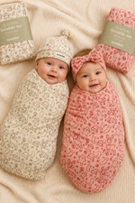 Two babies wrapped in patterned swaddles with matching hats and headbands on a beige blanket.