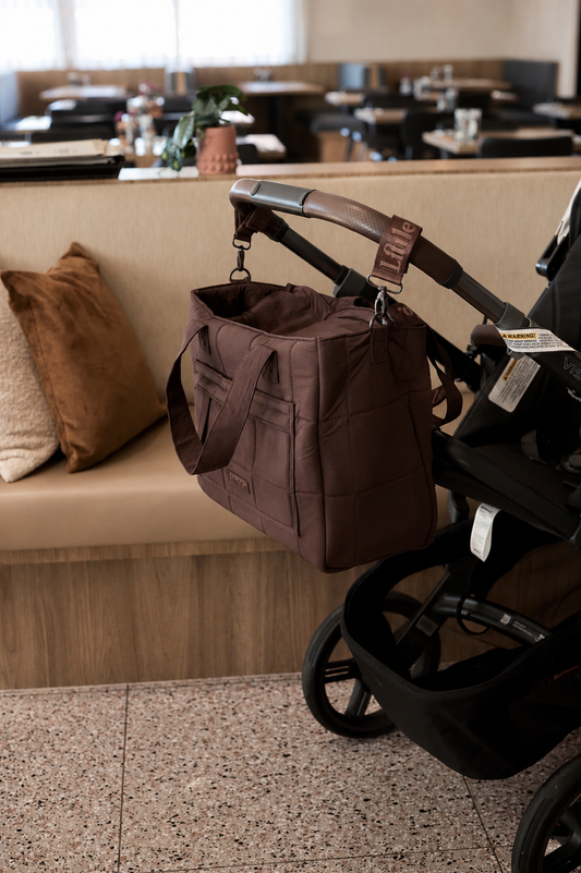 A brown quilted diaper bag hanging from a black stroller handle in a cafe setting.