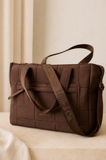 A brown quilted nylon tote bag with a matching adjustable shoulder strap, resting on a light beige stone surface against a neutral cream background.
