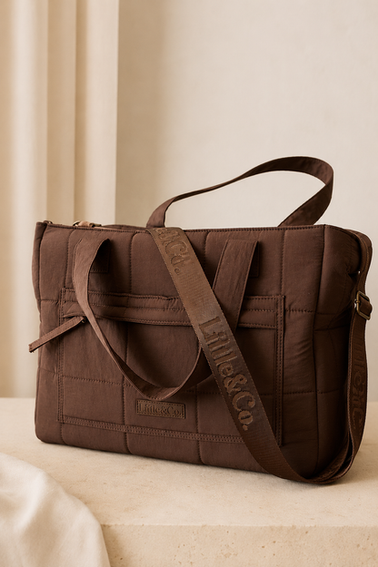 A brown quilted nylon tote bag with a matching adjustable shoulder strap, resting on a light beige stone surface against a neutral cream background.