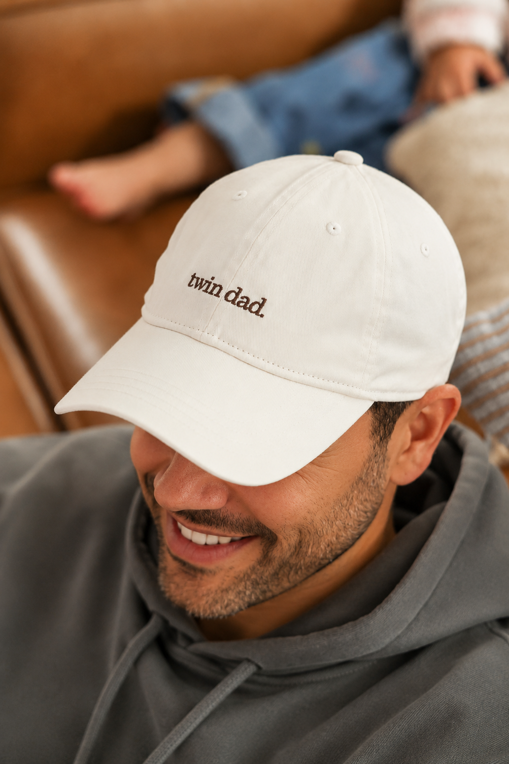 Man wearing a white cap with 'twin dad' text, sitting on a couch.