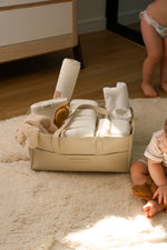 Leather Little & Co nappy caddy filled with baby essentials on a soft rug, with baby nearby in a warm nursery setting.