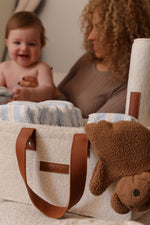 Cream bouclé nappy caddy with tan leather handles, filled with nappies and baby essentials, with a smiling baby and mum softly blurred in the background.