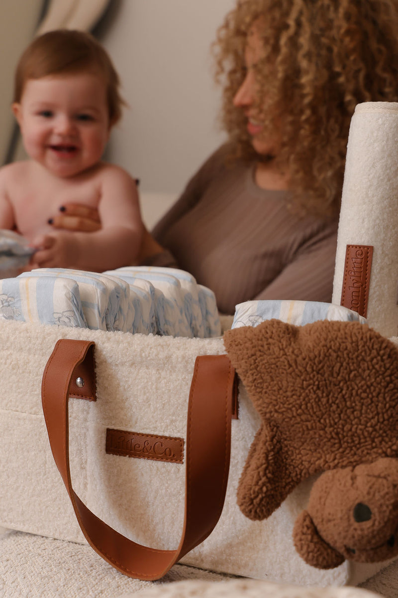 Cream bouclé nappy caddy with tan leather handles, filled with nappies and baby essentials, with a smiling baby and mum softly blurred in the background.
