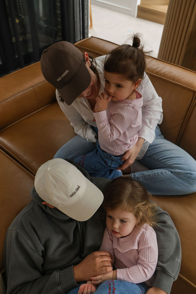 Two adults and two children sitting on a brown couch indoors. Parents are wearing twin mum and twin dad hat 