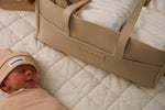 Beige leather Little & Co nappy caddy filled with baby essentials beside a newborn lying on a quilted mat.